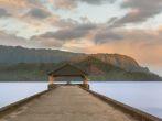 Rising sun illuminates the peaks of Na Pali mountains over the calm bay and Hanalei Pier in long exposure photo.